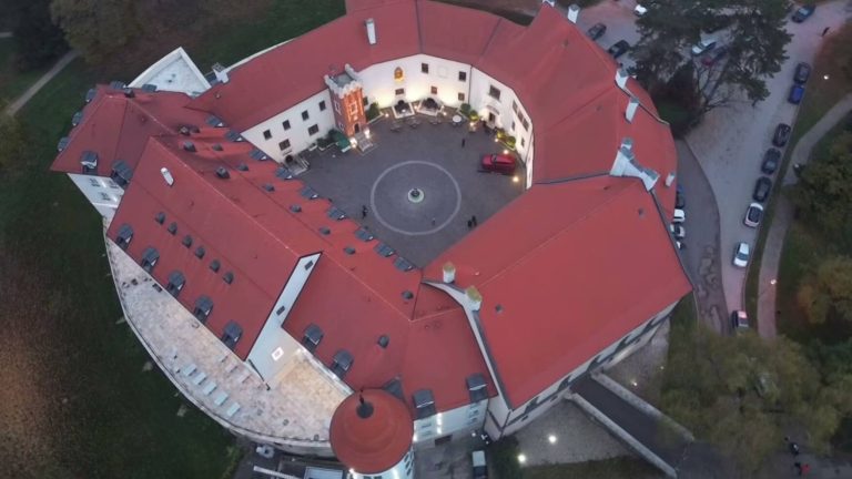An aerial view of a historic castle with a circular courtyard, illuminated at dusk, featuring red-tiled roofs and surrounding greenery.