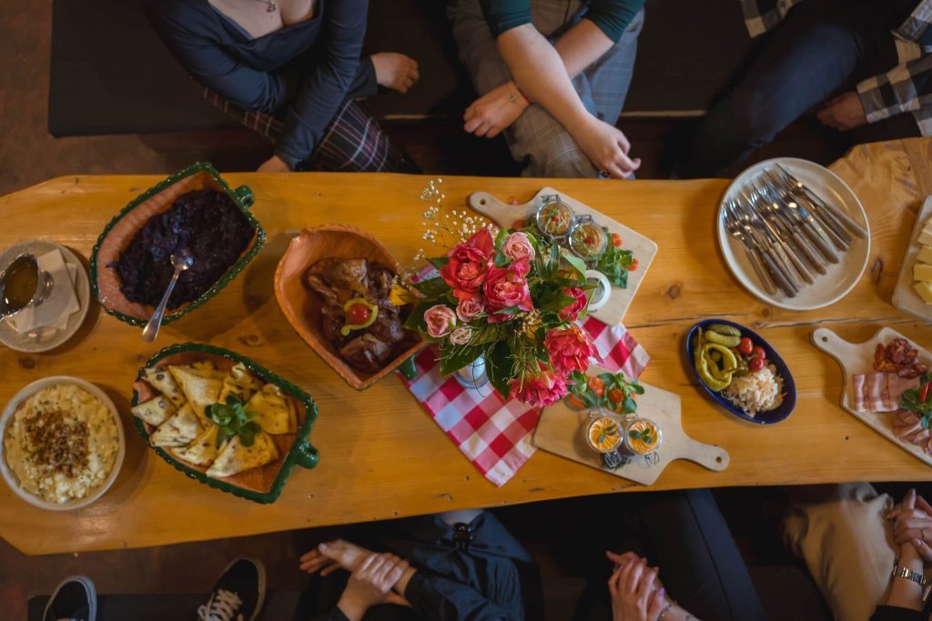 A cozy dining setting in Bratislava featuring a rustic wooden table filled with traditional Slovak dishes. The meal includes roasted meat, dumplings, cheese platters, pickled vegetables, and fresh salads, accompanied by a red and white checkered napkin and a bouquet of pink roses as the centerpiece. A group of people sits around the table, enjoying an authentic local culinary experience as part of an incentive nature getaway in Bratislava.