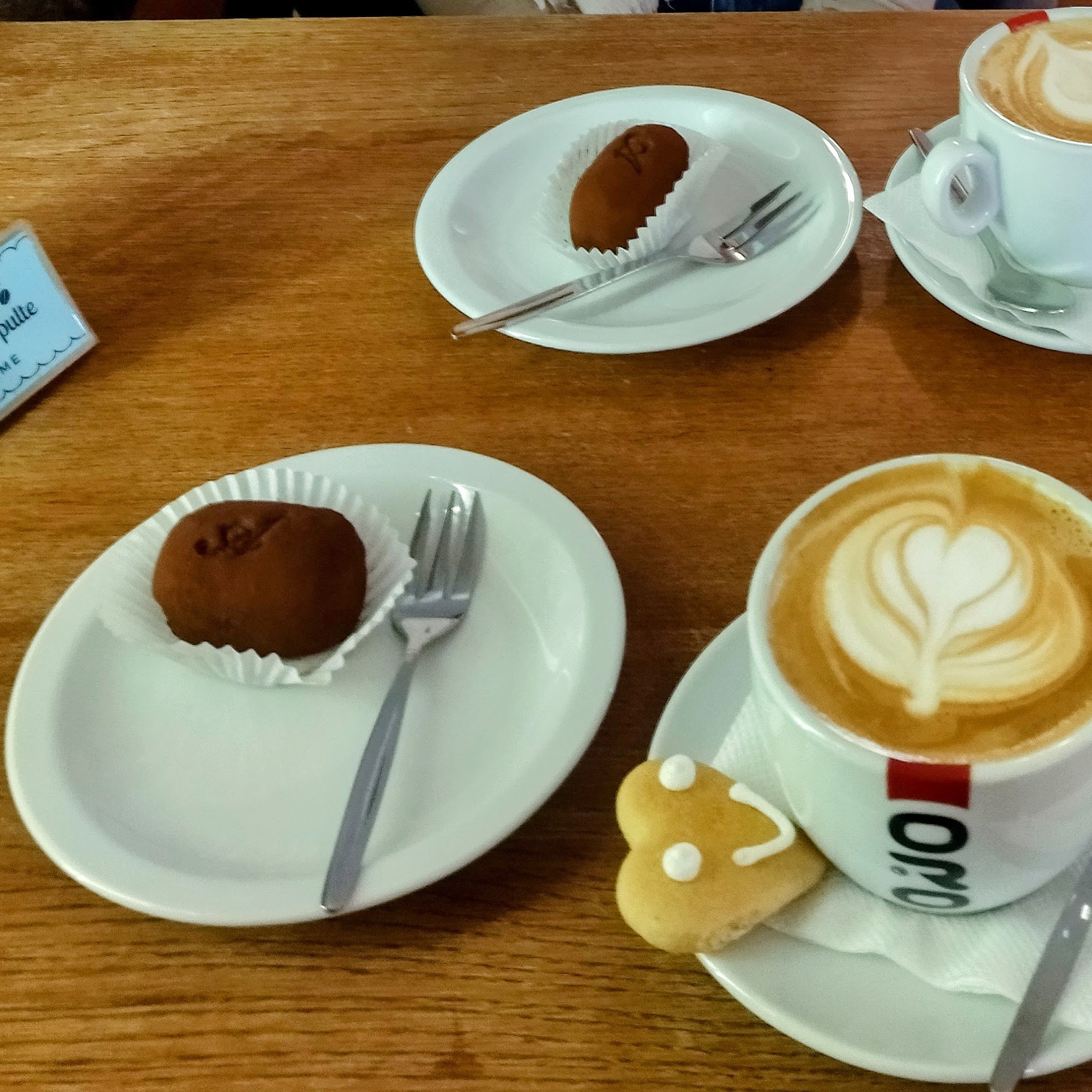 A cozy café setting featuring a cappuccino with heart-shaped latte art, accompanied by a smiling heart-shaped cookie. On the table, there are two servings of Kremnický Krumpel, a traditional handmade chocolate delicacy from Kremnica, Slovakia, with a soft, cocoa-dusted exterior. The warm wooden table and elegant white plates add to the inviting atmosphere
