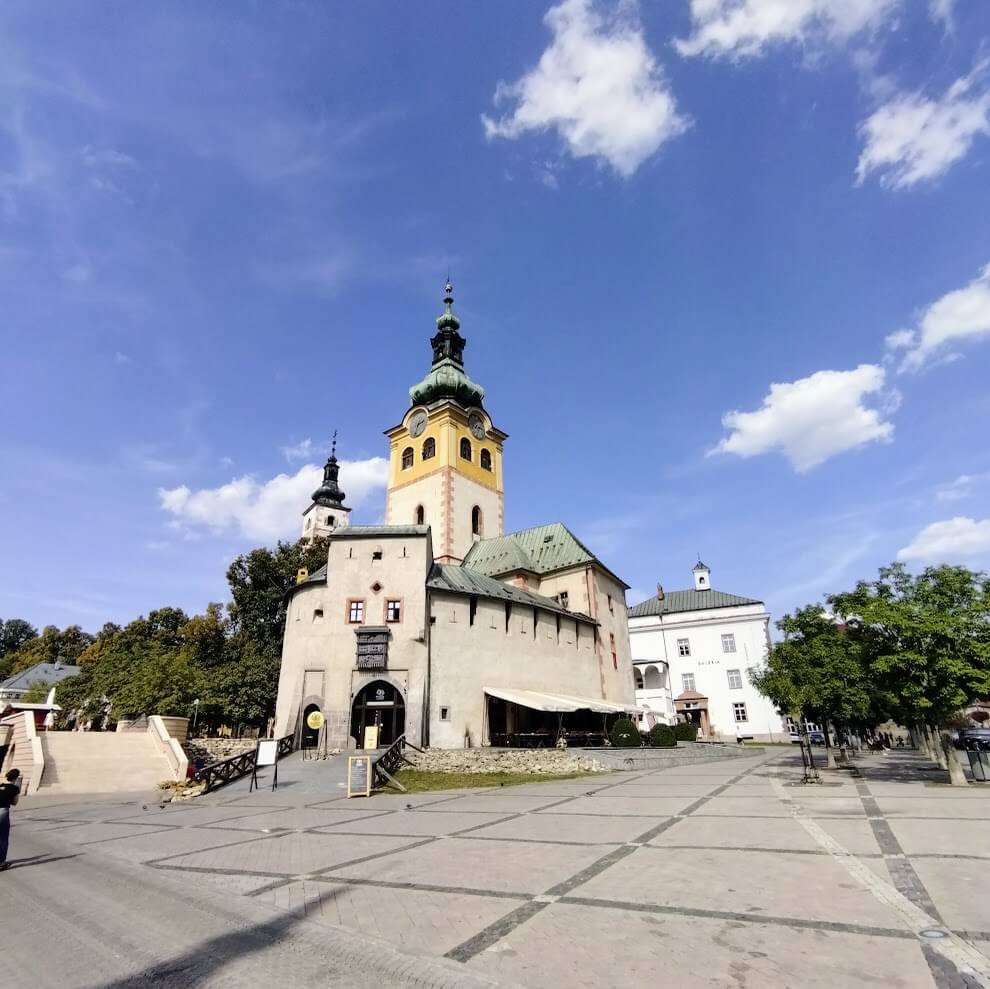 Historic Town Castle (Mestský hrad) complex in Banská Bystrica, Slovakia, featuring a prominent yellow bell tower with green copper dome, surrounded by connected buildings of various architectural styles. The complex stands on a plaza with patterned stone pavement, under a bright blue sky with scattered white clouds. Trees and outdoor seating areas are visible around the perimeter.
