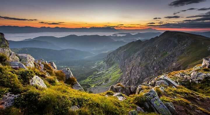 Image 1:
This image shows a breathtaking mountain landscape at sunset or sunrise. The view is captured from a high vantage point on a rocky mountain peak with moss and small vegetation growing between the rocks in the foreground. The scene overlooks a deep valley with dramatic mountainous terrain stretching to the horizon. Multiple layers of mountains fade into the distance, creating a beautiful blue-purple haze effect. The sky displays vibrant sunset/sunrise colors with orange and pink hues along the horizon, transitioning to darker blue above with some scattered clouds. This appears to be a view from one of the mountain ranges in Slovakia or another Central European country, possibly the Tatras or Carpathian Mountains, showcasing the natural beauty of the region's alpine landscape.