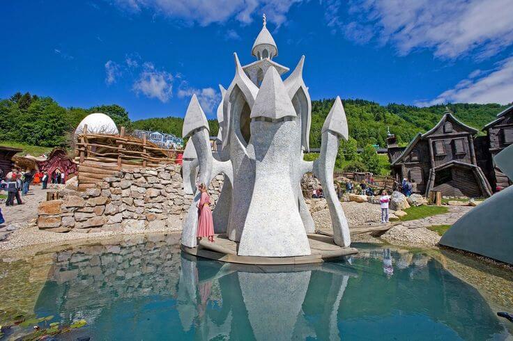 This image shows an unusual modern sculpture or architectural structure in what appears to be a theme park or cultural attraction. The central feature is a striking white stone or concrete structure with a distinctive design featuring pointed, angular elements that create a stylized castle or fantasy-like appearance. The structure sits in a shallow reflecting pool that mirrors its image. Around this central piece are other themed elements including what looks like rustic wooden structures or buildings. Visitors can be seen walking around the attraction, and the setting includes stone walls and landscaped areas. The scene is set against a backdrop of forested hills under a blue sky with scattered clouds. This appears to be some kind of cultural park or themed attraction, possibly representing Slovakian folklore or fantasy elements.