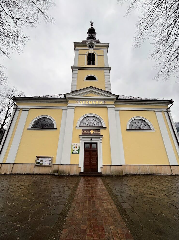 This image shows the facade of a yellow church or basilica with white architectural trim and details. The building features a prominent bell tower with a black dome and a classical architectural style. The facade has semi-circular windows above a central wooden door, and "AVE MARIA" text is visible on the front of the building. A brick pathway leads directly to the entrance, and the photo is taken during winter or early spring, as the surrounding trees are bare. The sky is overcast, giving the scene a somber atmosphere. There appears to be an information board or display to the left of the entrance. The building style suggests it's likely a Catholic church in Central or Eastern Europe.