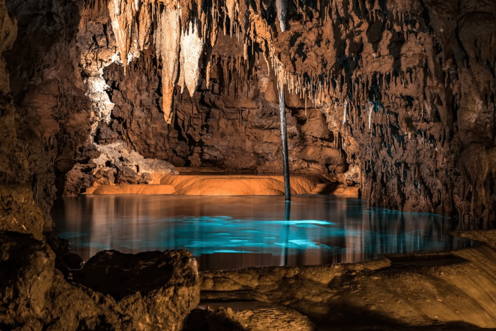 This image shows a stunning underground cave formation with dramatic stalactites hanging from the ceiling. The cave features a striking turquoise blue underground pool or lake with crystal clear water. The rock formations appear to be limestone in various orange-brown tones, illuminated by what must be artificial lighting to highlight the natural features. A thin pole or support column can be seen extending down into the water. The serene water surface perfectly reflects the surrounding cave formations, creating a mirror-like effect. This is likely part of a cave system open to tourists, possibly in Slovakia or another karst region of Central Europe.