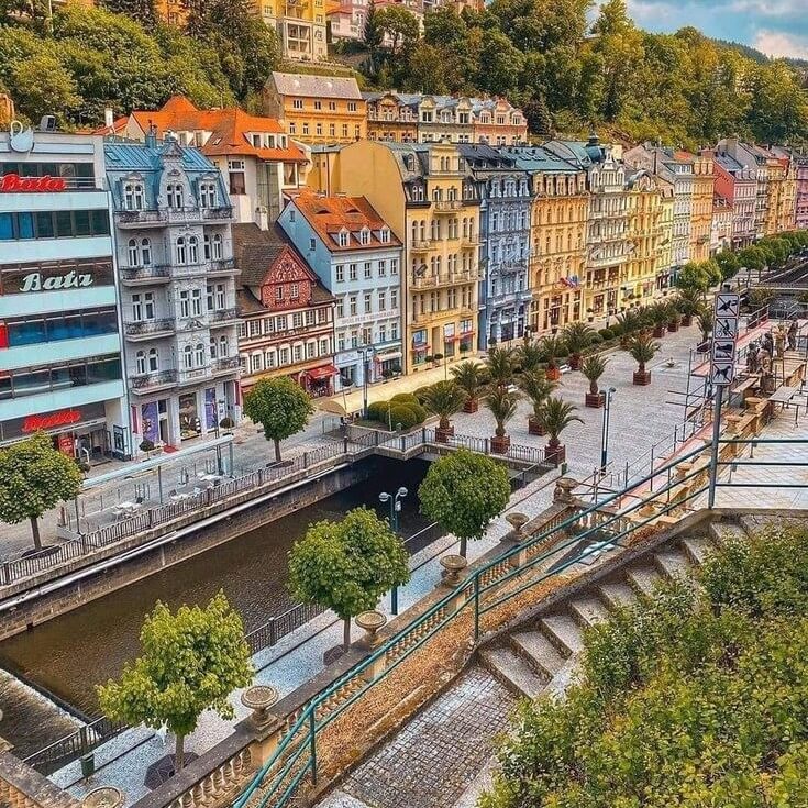 A vibrant European street scene in Karlovy Vary, Czech Republic, showcasing a row of colorful historic buildings along a canal lined with trimmed trees and palm planters. Stone steps lead down to the canal, and lush greenery covers the hillside in the background during Central Europe Itinerary Tour