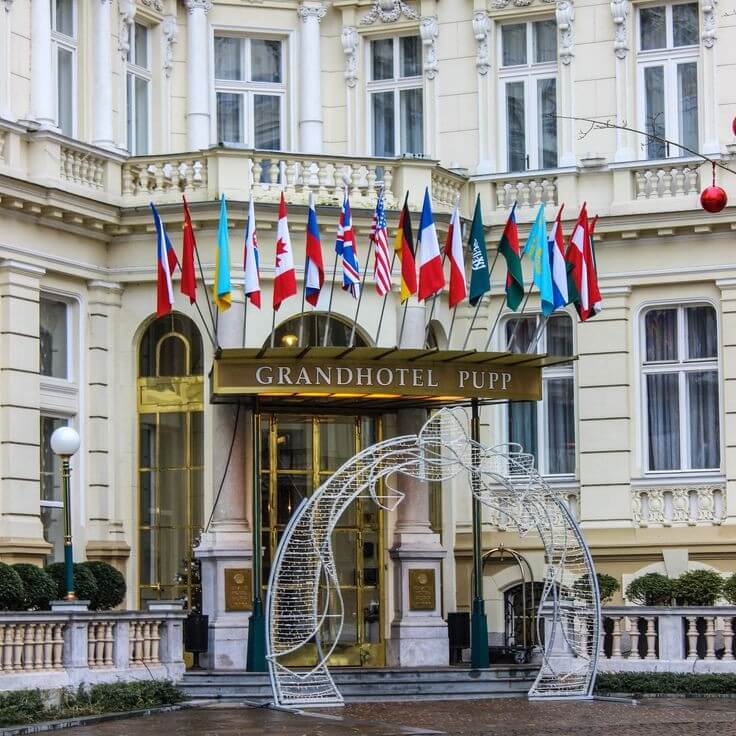 The elegant entrance of the Grandhotel Pupp in Karlovy Vary, Czech Republic. The building features classic European architecture with ornate details, multiple national flags above the entrance, and a decorative arch structure in front of the golden glass doors