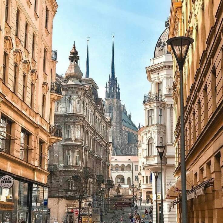 View of a historic street in Brno, Czech Republic, with elegant classical buildings lining both sides and the twin spires of the Cathedral of St. Peter and Paul visible in the background against a clear blue sky.