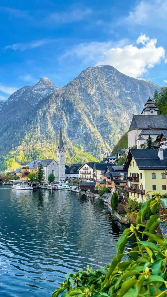 A breathtaking view of Hallstatt, Austria, with its picturesque village nestled between a clear lake and towering Alpine mountains. The scene features charming traditional Austrian buildings including a church with a tall spire, colorful houses along the waterfront, and small boats docked at the shore. The dramatic mountain backdrop rises steeply behind the village with some trees visible on the lower slopes, all under a bright blue sky with scattered clouds.