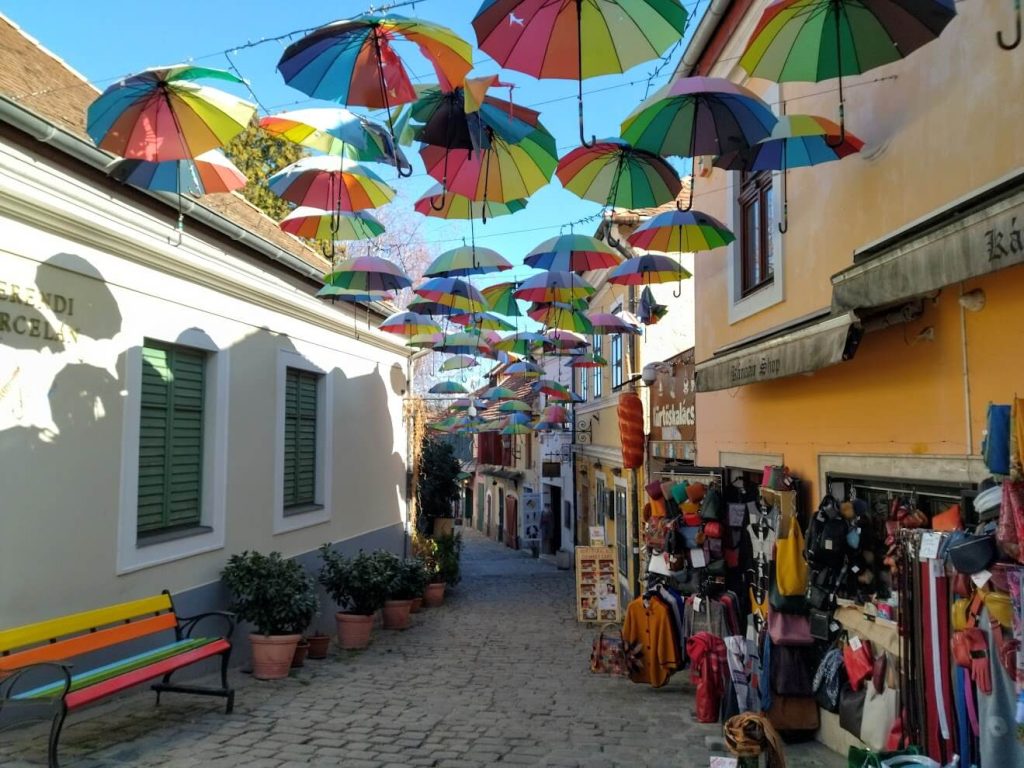 A charming cobblestone street in Szentendre, Hungary, decorated with colorful rainbow umbrellas suspended overhead between buildings. The narrow pedestrian lane features pastel-colored buildings with a rainbow-painted bench on the left, potted plants along the street, and a shop selling bags and souvenirs on the right. The installation of hanging umbrellas creates a whimsical, artistic atmosphere typical of this cultural town near Budapest you will discover during Central European Itinery