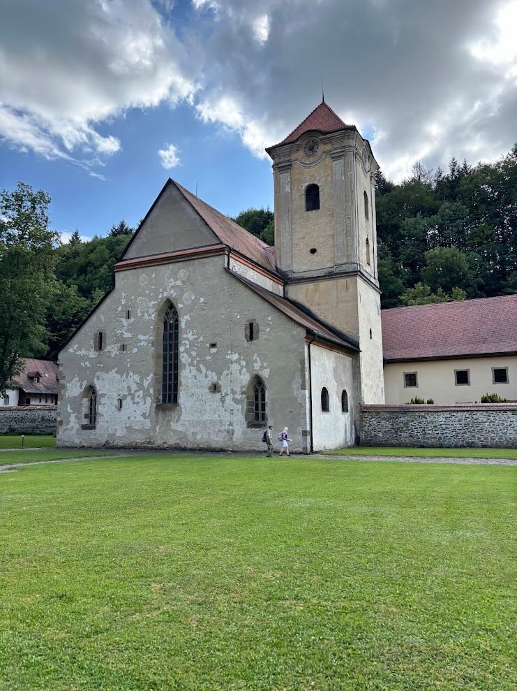 Historic Red Monastery (Červený Kláštor) in Slovakia, featuring a tall beige church building with a red-tiled roof and Gothic-style arched windows, set against a backdrop of trees and cloudy sky, with two visitors walking nearby on the green lawn.