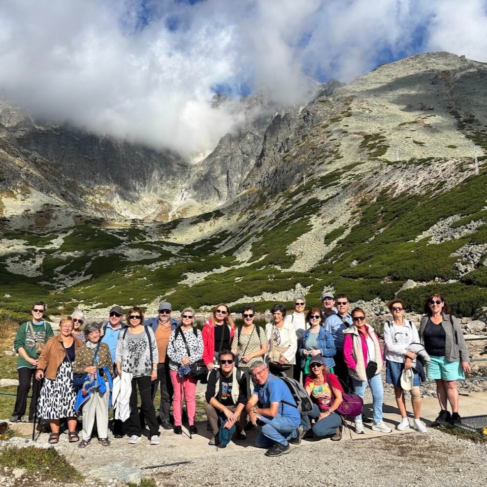 A group of international travelers posing together in the High Tatras mountains of Slovakia, with dramatic rocky peaks and clouds in the background during a guided tour.