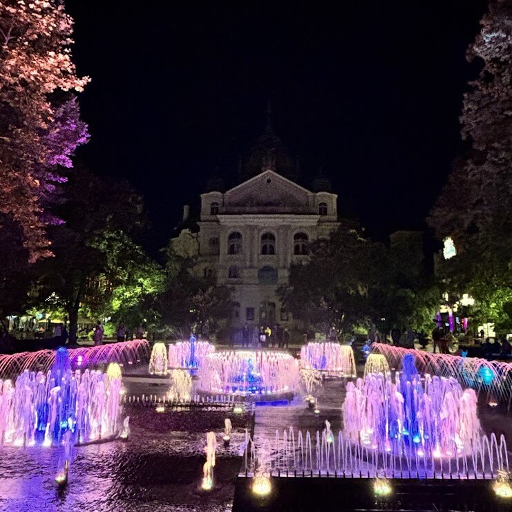 Illuminated singing fountain in Kosice at night with colorful purple and blue lights and historic building in background"