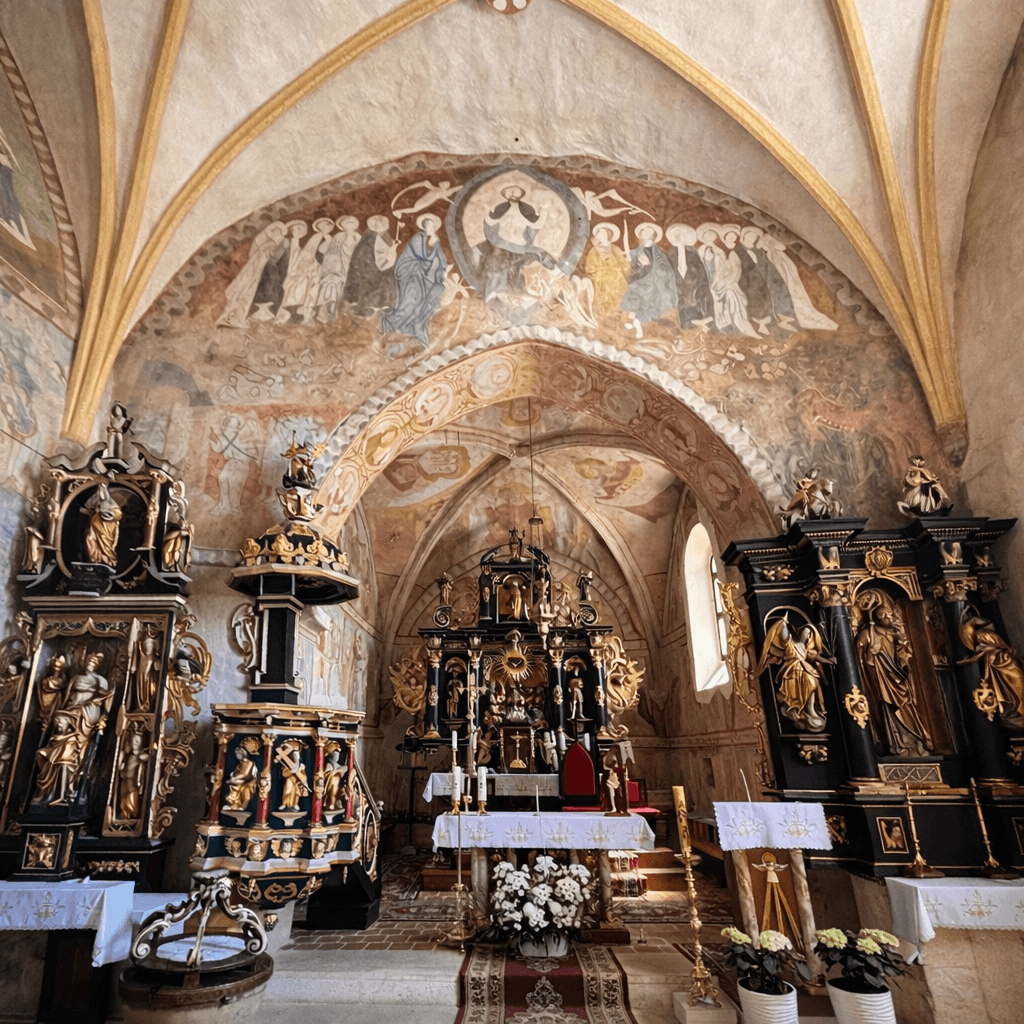 Interior of the Church of the Holy Spirit in Žehra, Slovakia, showing Gothic frescoes covering the vaulted ceiling and arches, with a Baroque altar, wooden pulpit and side altars decorated with gilded sculptures in the foreground.