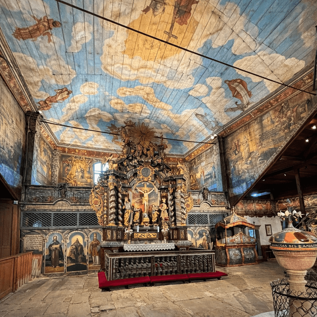 Interior of the Wooden Articular Church in Kežmarok, Slovakia, showing a richly painted wooden barrel-vaulted ceiling depicting clouds and biblical scenes, a gilded Baroque altar with a Crucifixion sculpture, colourful painted walls, stone floor and a decorative baptismal font in the foreground.